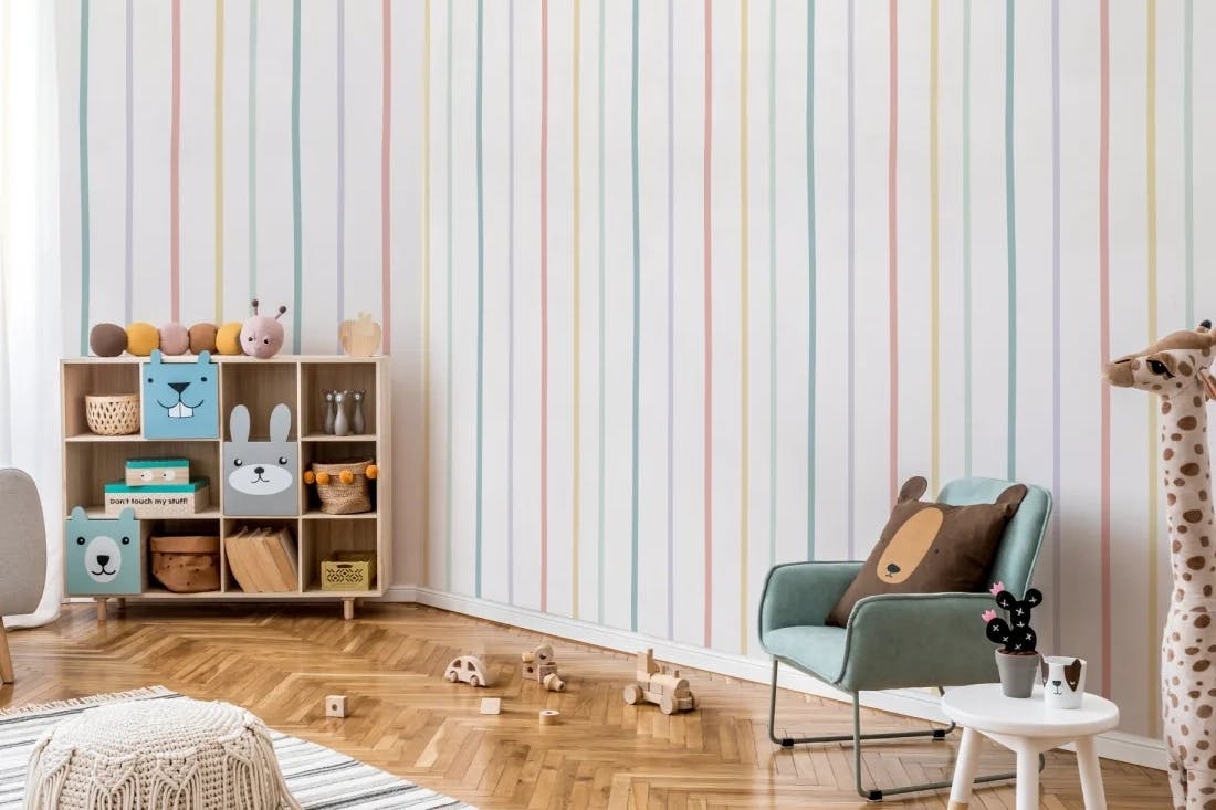 Nursery with wallpaper in white with narrow individual bands of rainbow colours, parquet wood floor with a white rug with thin bands of blue, storage unit with animal fronts to sections, filled with baskets and boxes, blue upholstered child's chair with bear cushion, wooden toys on floor, giraffe plush by small white painted wooden round side table. 