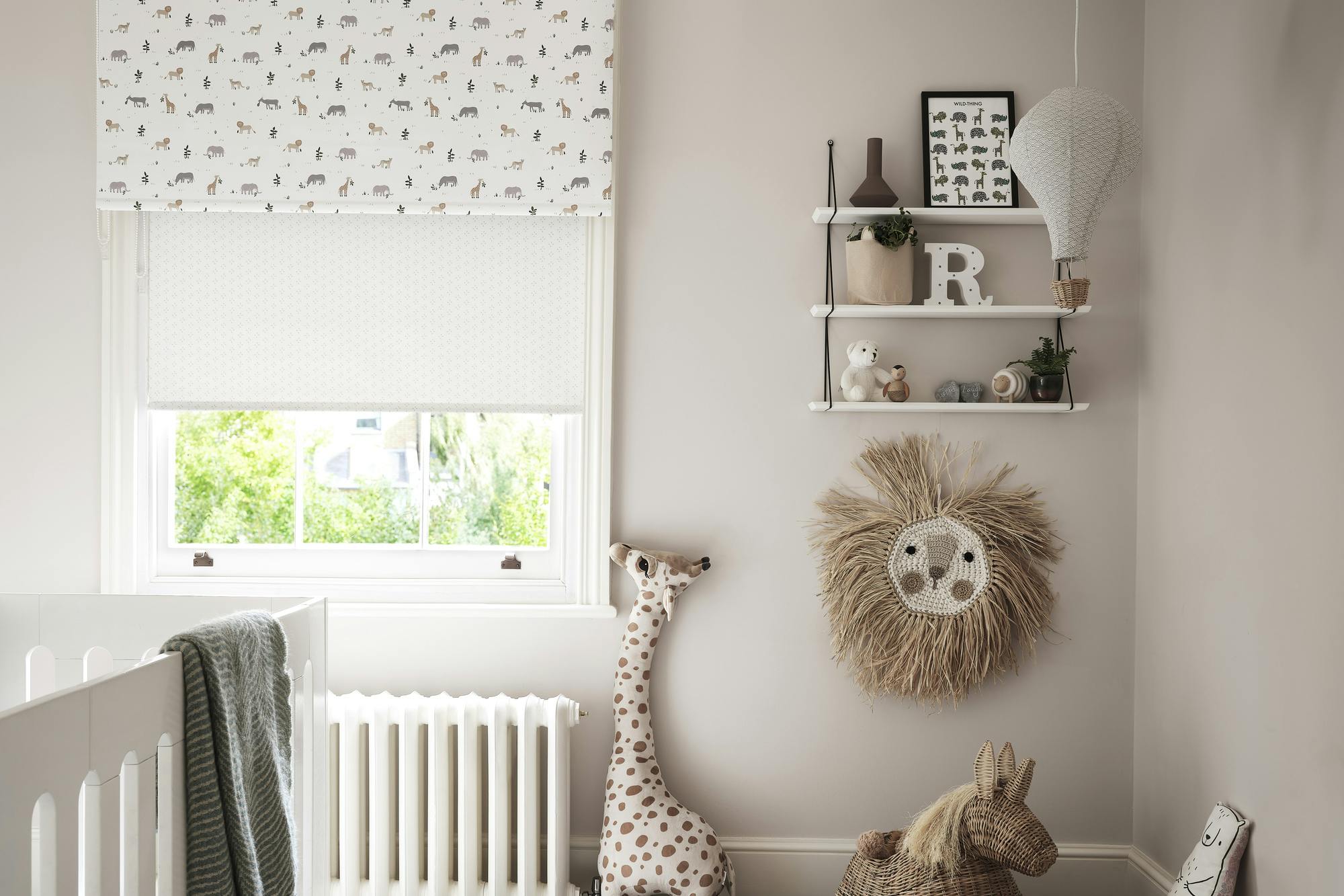 Child's bedroom with natural walls and sash window dressed in two layered roller blinds, neutral with tiny yellow bursts behind a white with colourful jungle animals, wooden floor with a round faded-effect rug in white and voilet, white cot, jungle-theme animals in corner. 