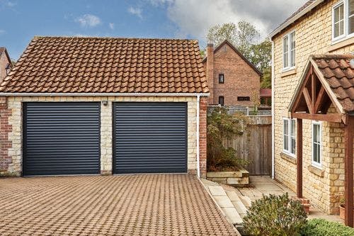 Detached stone garage with two dark grey roller doors and a tiled roof, next to a brick driveway and traditional-style house.