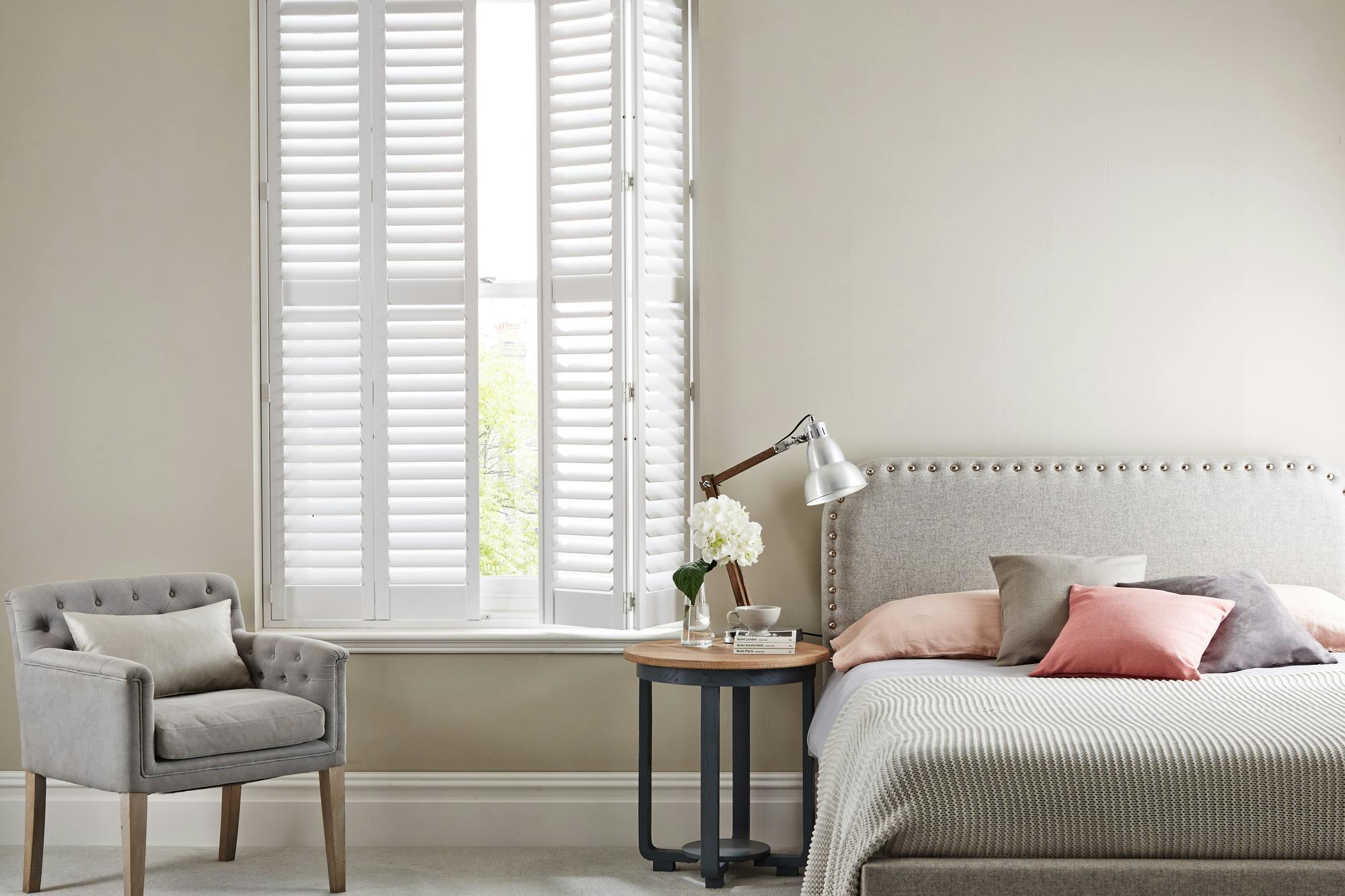 Neutral bedroom with tall window dressed in white shutters, right hand partially open, pale mink upholstered bedhead with pinstriped mink and white bedding, cushions in mink and dusky peach, mink upholstered chair, round bedside table with anglepoise lamp and two white blooms in small vase. 