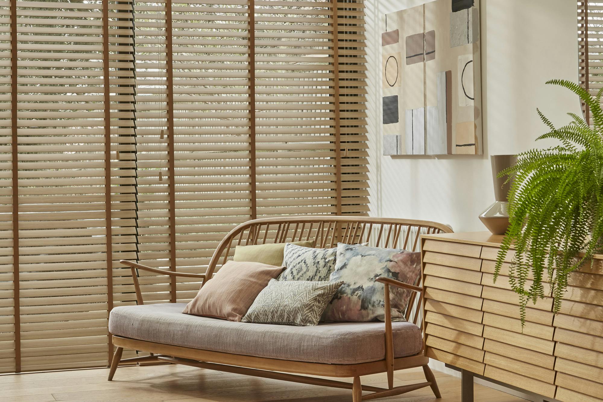 Living room in cream with windows and doors dressed in faux wood mid colour venetian blinds, fully lowered and tilted, low wooden sofa with cushions in pale peach and watercolour-effect patterns, wooden side unit with large fern, wooden floor with rug in cream with black dashes, light rays on walls. 