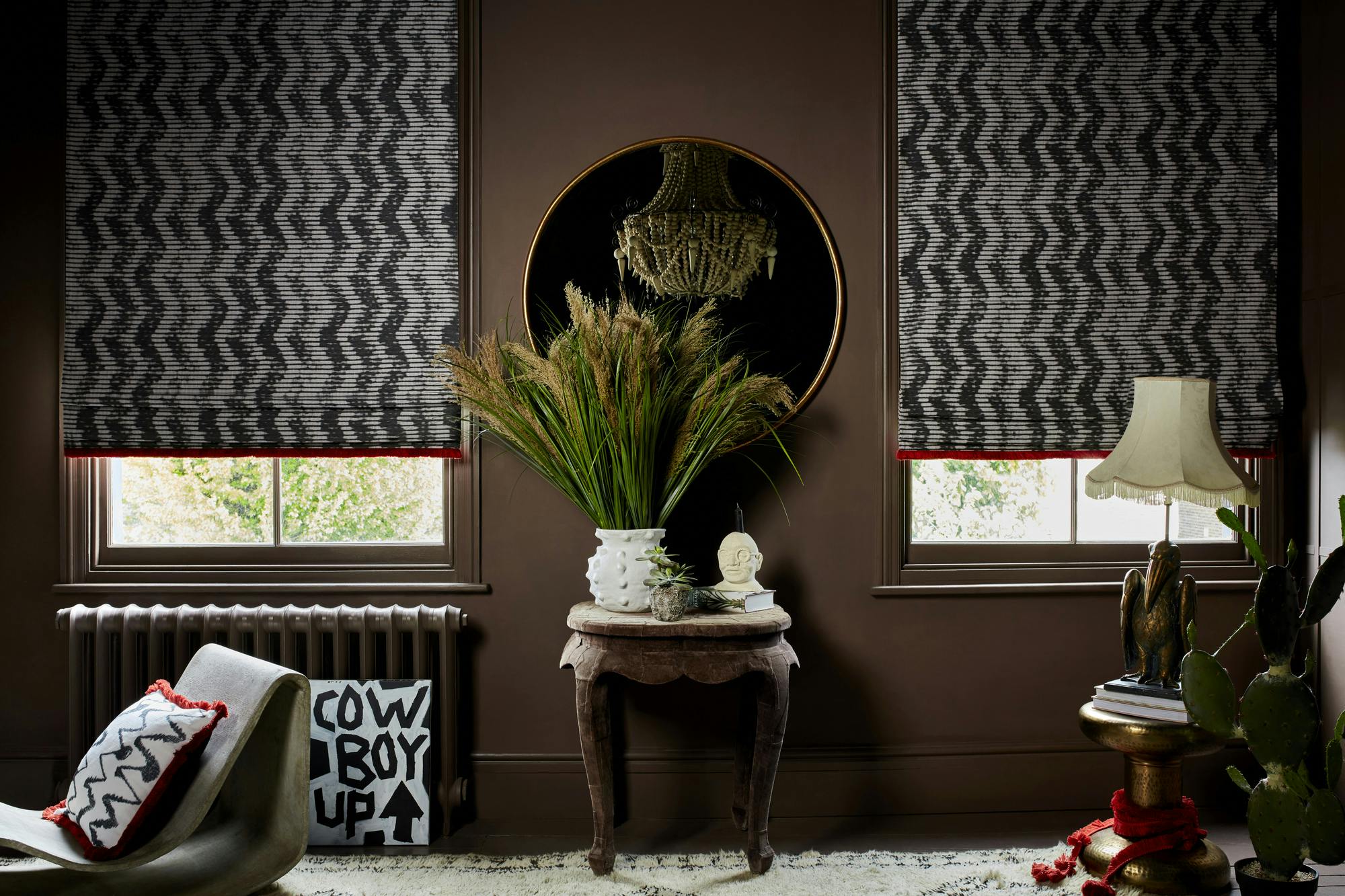 Close in on room with dark brown walls, two windows dressed in pale grey Roman blinds with a vertical soft zigzag pattern in charcoal, with an orange frill trim three quarters lowered, small side table between windows with white pot filled with pampas grass, to right a small round side table with lamp with cream shade with frill, on left a saddle chair with cushion matching the blinds.