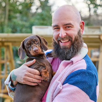 Influencer Luke Newnes, wearing a pink and blue fleece in his garden holding a dark brown daschund.