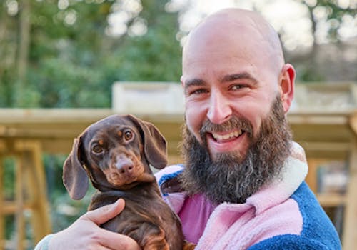 Influencer Luke Newnes, wearing a pink and blue fleece in his garden holding a dark brown daschund.