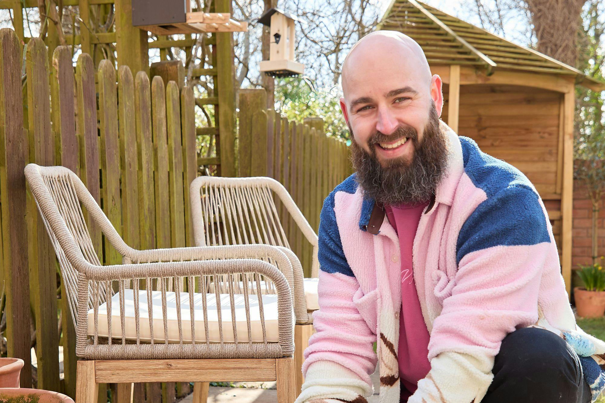 Influencer Luke Newnes, wearing a pink and blue fleece in his garden planting purple and yellow flowers in a pot with multiple pots and shoots around on a sandstone-style patio next to two raffia chairs with cream cushions.