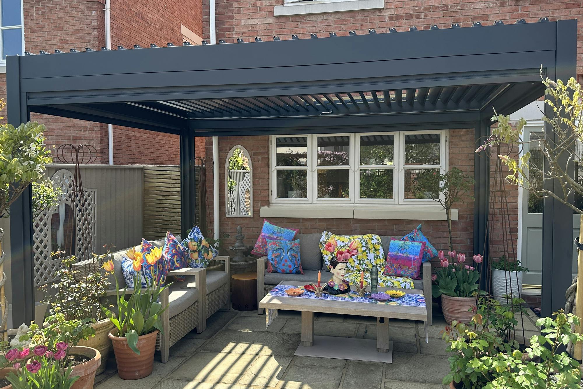 Exterior of new build house with sandstone-style patio under a grey freestanding pergola, two wooden sofas with grey cushions, and colourful cushions, low table with colourful decorations, multiple pots with flowers, shrubs and trees.