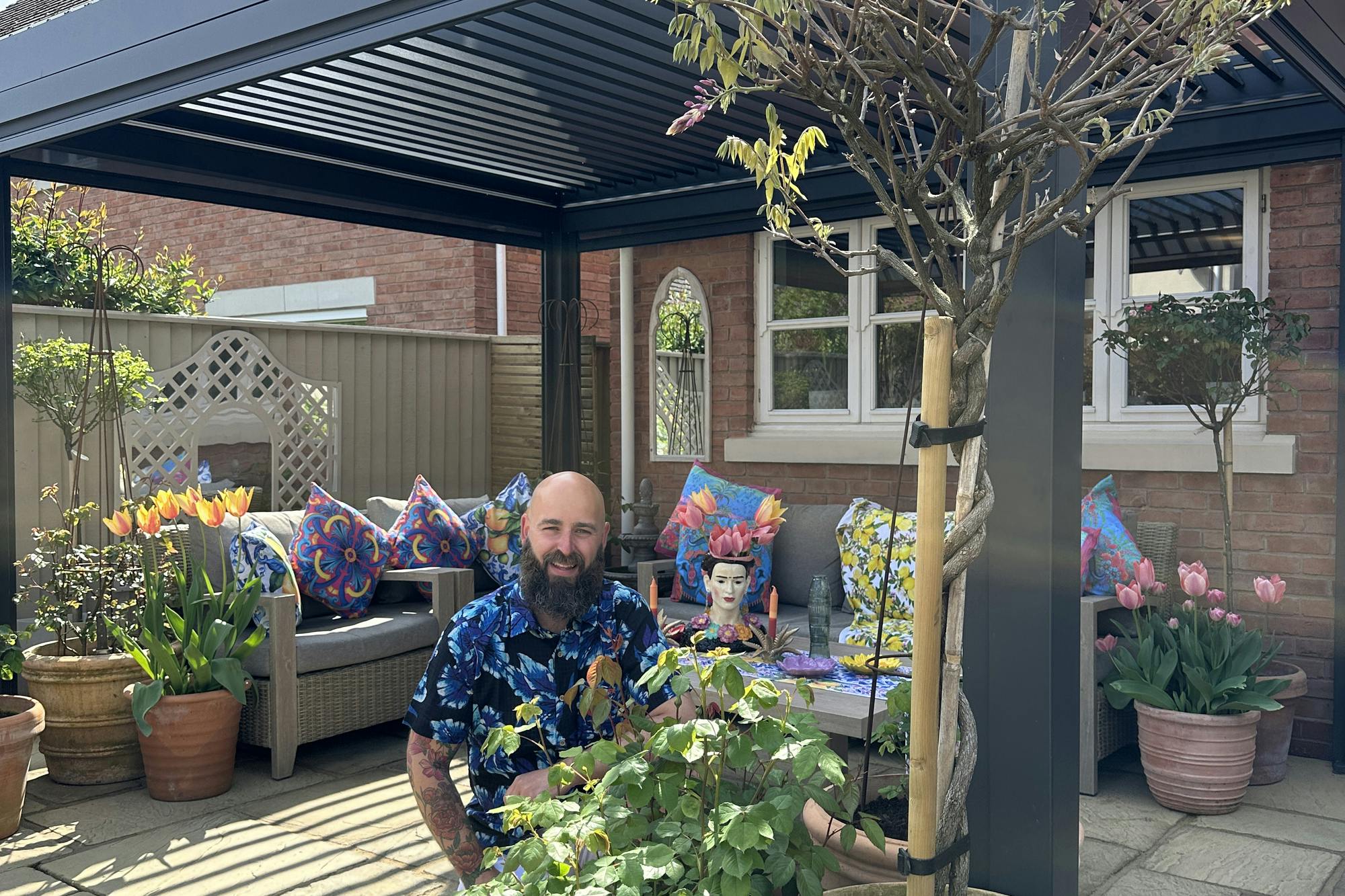 Exterior of a new build property, on a laid sandstone-style patio, a man in a hawian shirt kneels beside a tall tree in a large terracotta pot, with a smaller shrub in a terracotta pot, in front of a grey fresstanding pergola, with pots of flowers and trees and a grey sofa with colourful cushions.