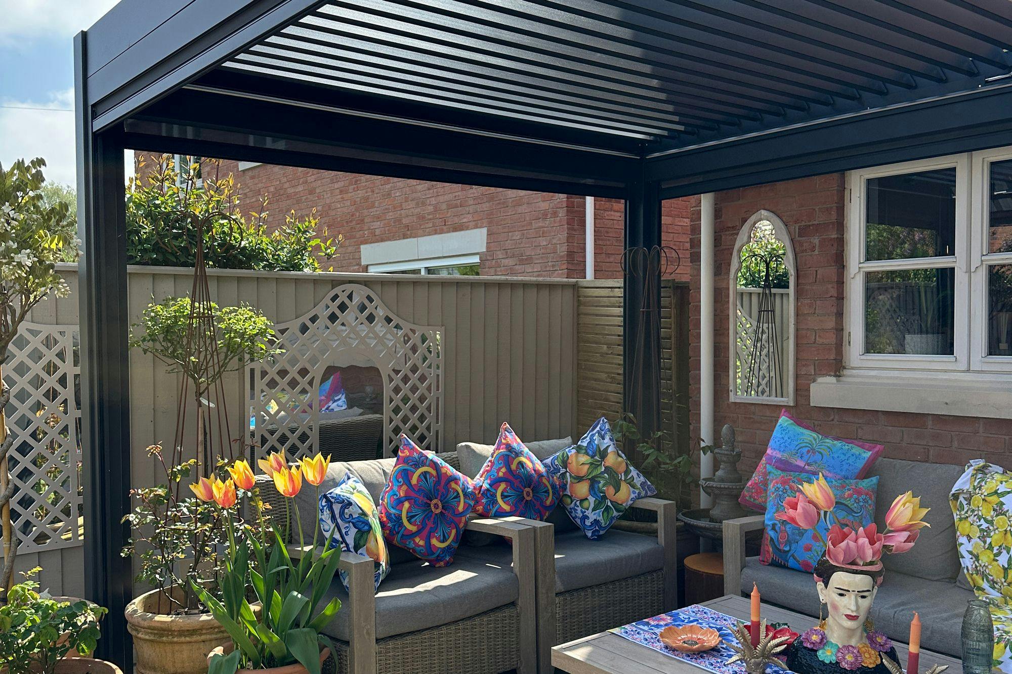 Exterior of new build house with sandstone-style patio under a grey freestanding pergola, two wooden sofas with grey cushions, and colourful cushions, low table with colourful decorations, multiple pots with flowers, shrubs and trees.