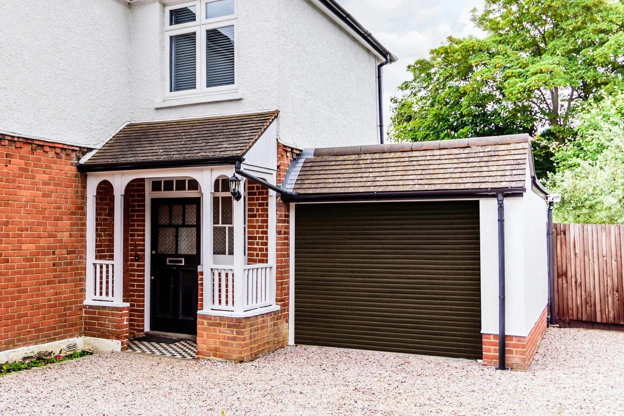 Corner shot of two storey house in reddish-brown brick to lower and white render to upper with dark brown roller garage door on garage attached to right, matching front door with white porchway railings and facia and shingled driveway.