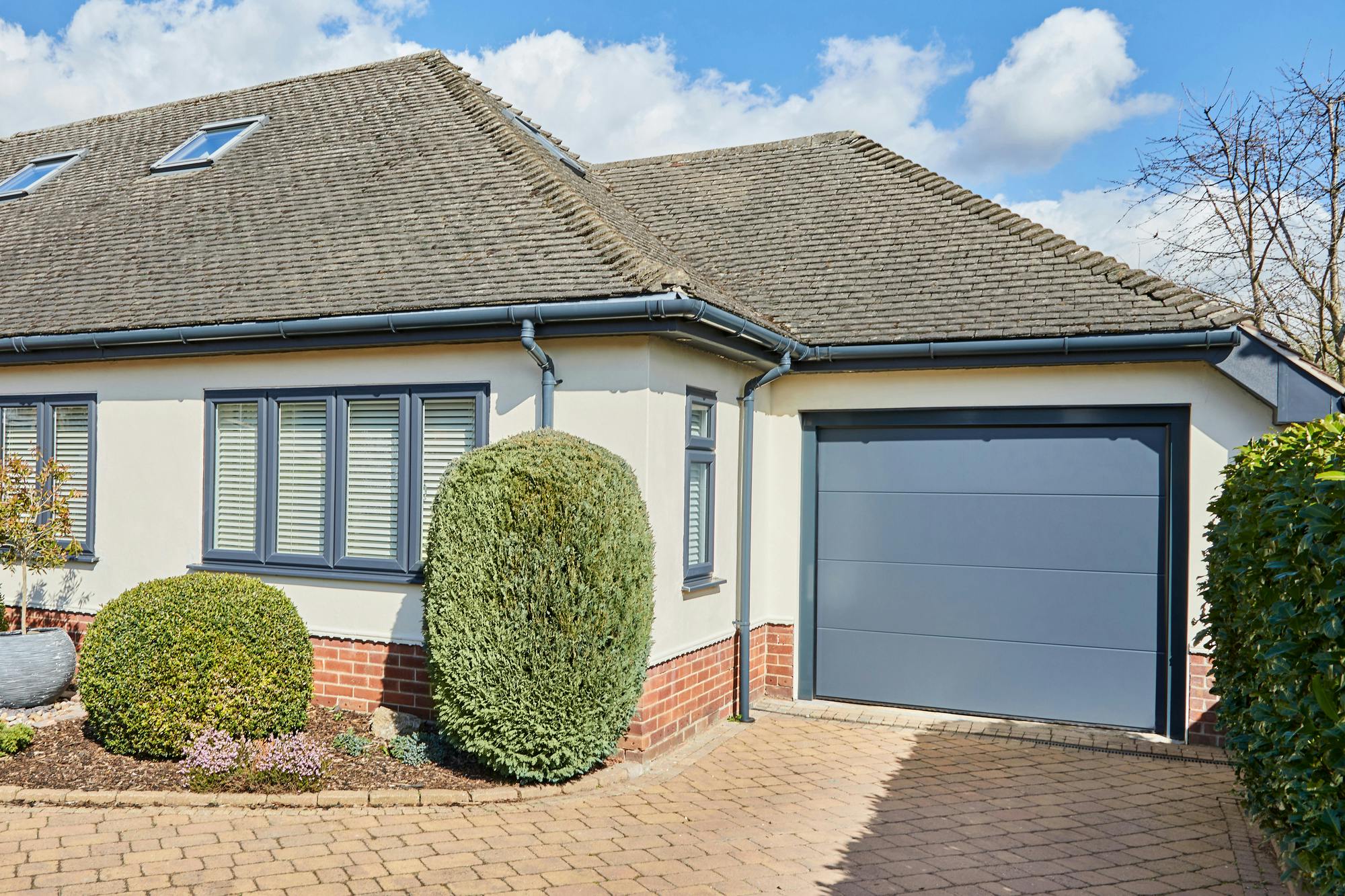 Corner shot of single storey house in white with anthracite grey integral sectional garage door matching guttering and window frames and block paved driveway.