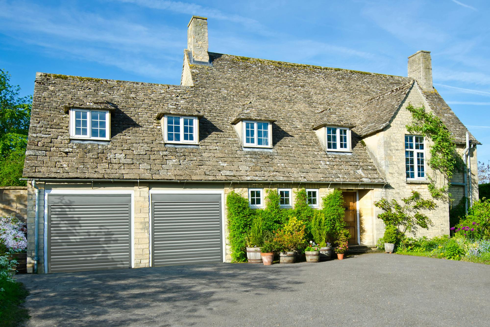 Large traditional pale stone house with wide driveway and integral double garage on left with two grey garage doors.