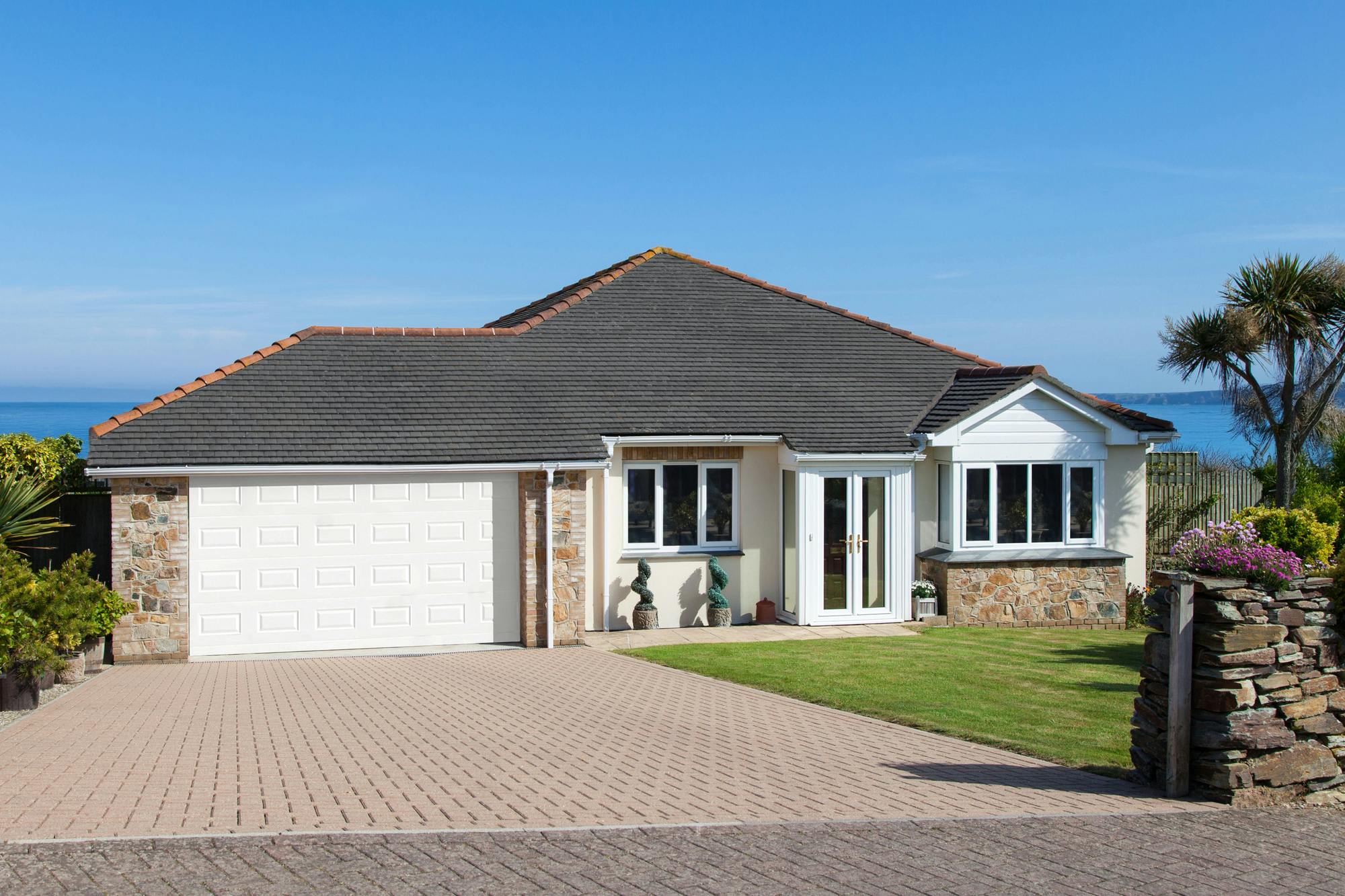 Exterior shot of modern brown-brick bungalow with integral garage door with georgian-effect panelling in white, closed, matching front door, white fascias to windows, brick driveway with lawn to right and flower pots to left of drive.