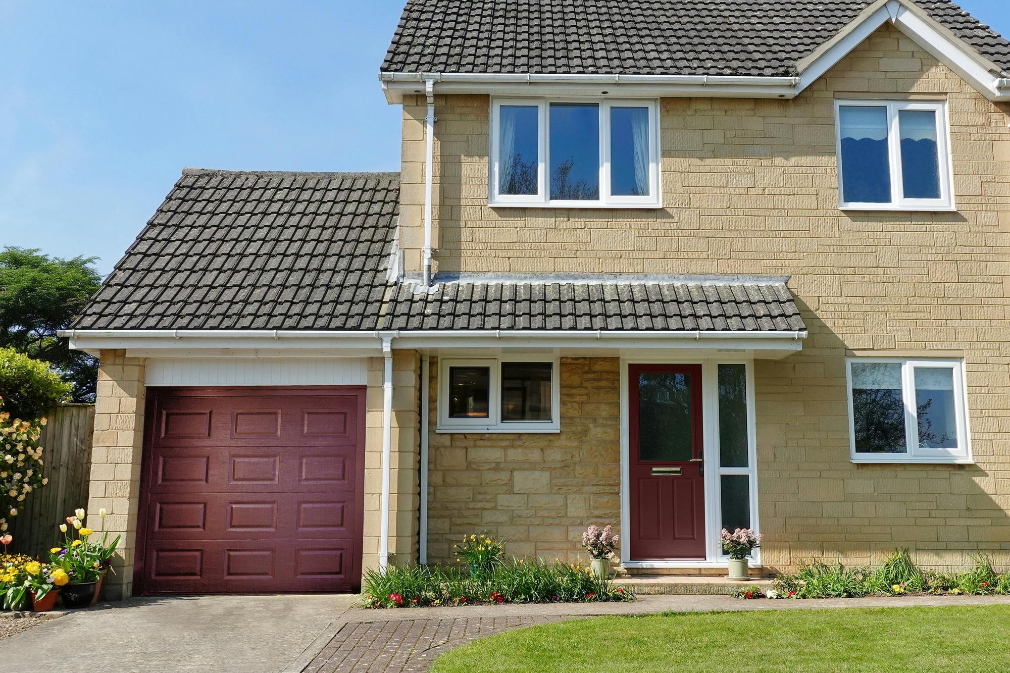 Exterior shot of modern brown-brick house with integral garage door in burgundy with georgian panel effect, closed, matching front door, white fascias to two sets of windows on ground floor, two windows above, asphalt driveway with lawn to right and flower borders to front of house and left of garage..