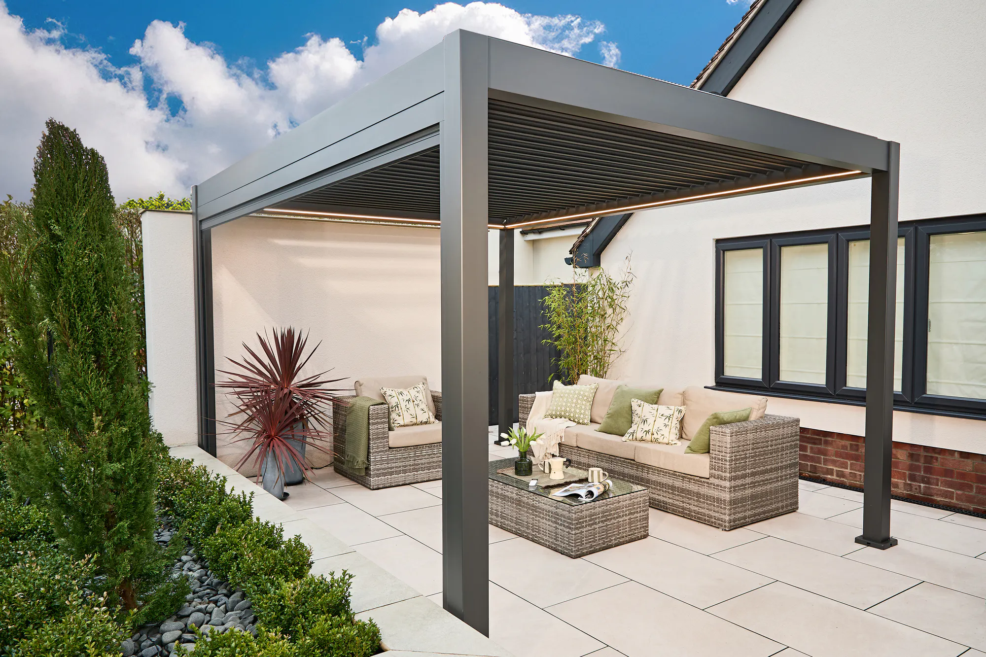 Exterior in a garden of a freestanding pergola in grey, installed on a cream patio next to a cream rendered house and a raised flower border with alpine plants and a raffia style sofa and coffee table under the pergola.