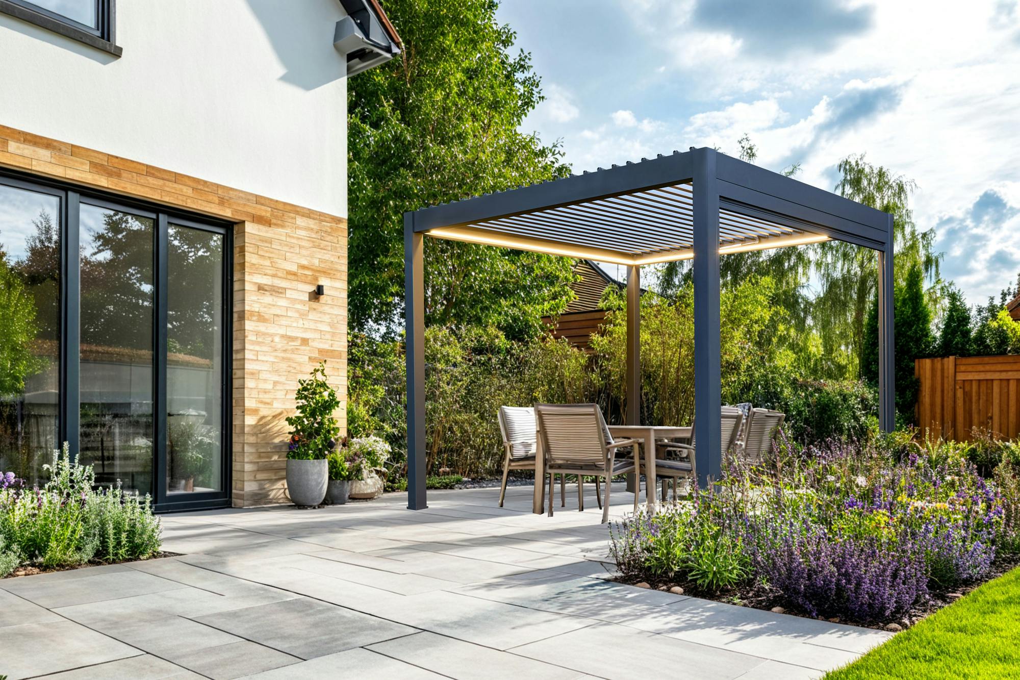 Exterior of a modern house, with a large paved patio down to a lawn with small inset flower beds at random points in the patio and a freestanding grey pergola with louvred roof over four chairs and a table,