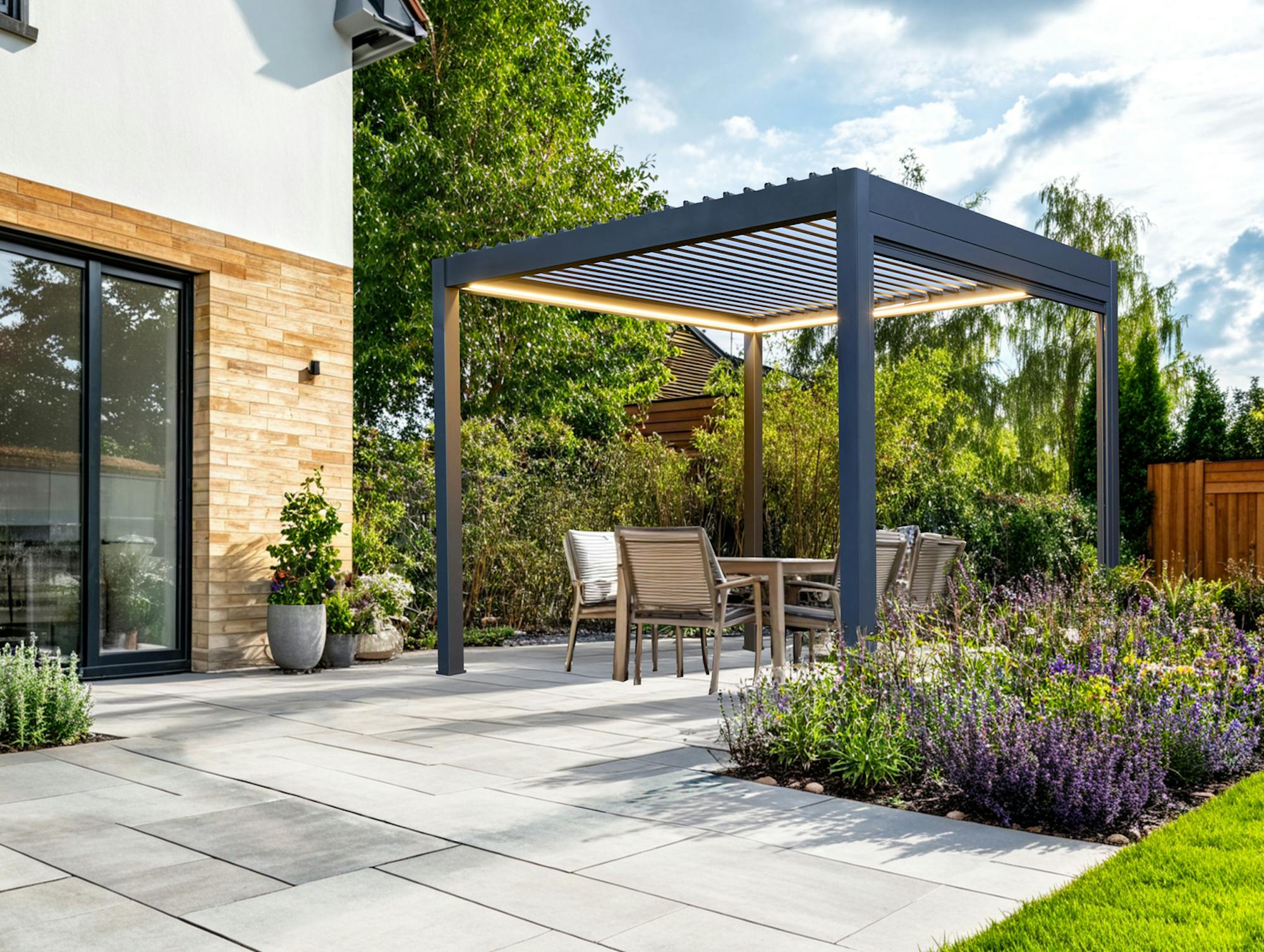 Exterior of a modern house, with a large paved patio down to a lawn with small inset flower beds at random points in the patio and a freestanding grey pergola with fabric roof over four chairs and a table.