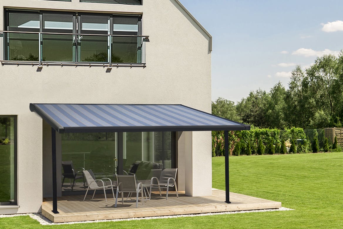 Large modern house and lawned garden with a pergola awning in mid blue and grey stripes extended over a pale wooden deck with modern furniture.