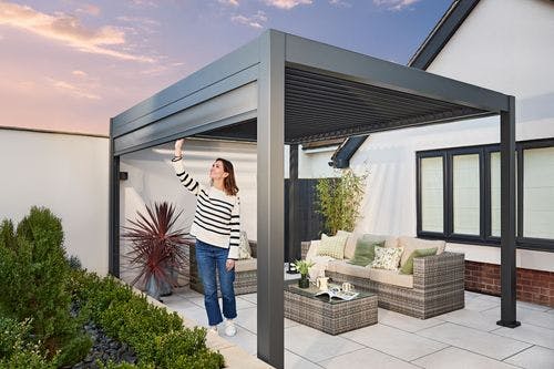 Woman adjusting a modern grey pergola in a stylish patio area with rattan furniture, greenery, and tiled flooring at sunset.