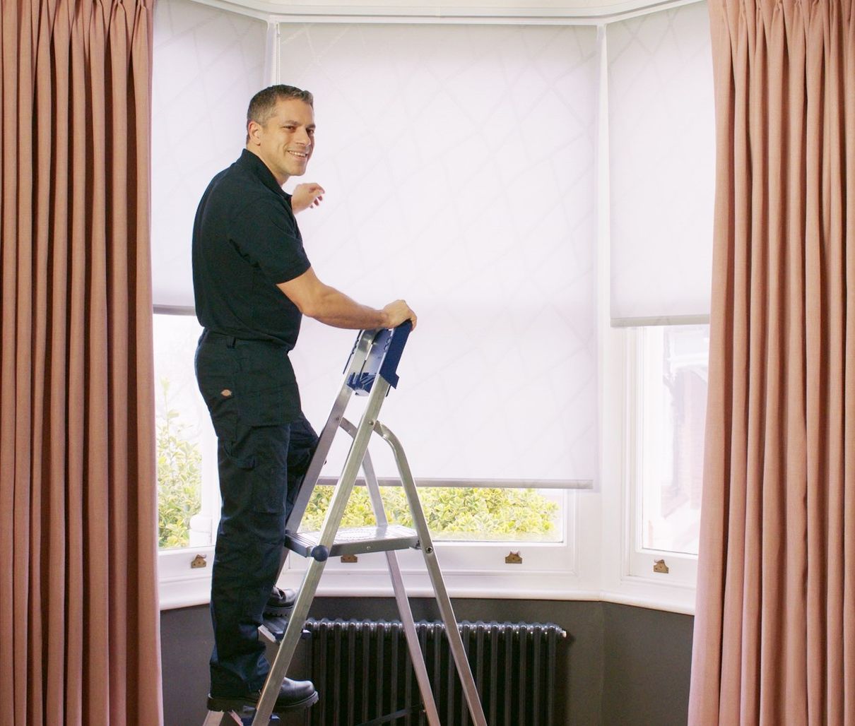 Hillarys advisor wearing branded uniform, fitting white roller blinds on a window, assisting customers in their home. 