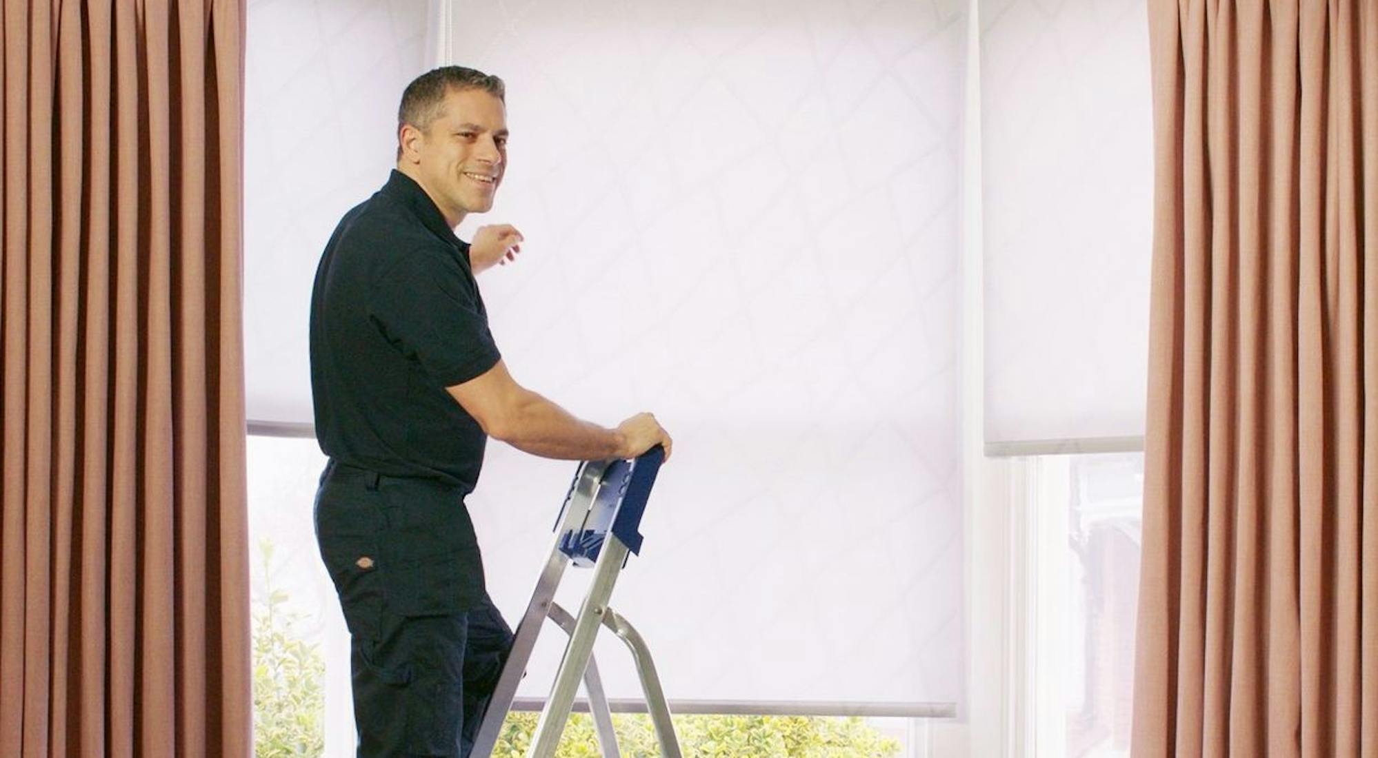 Hillarys advisor wearing branded uniform, fitting white roller blinds on a window, assisting customers in their home.