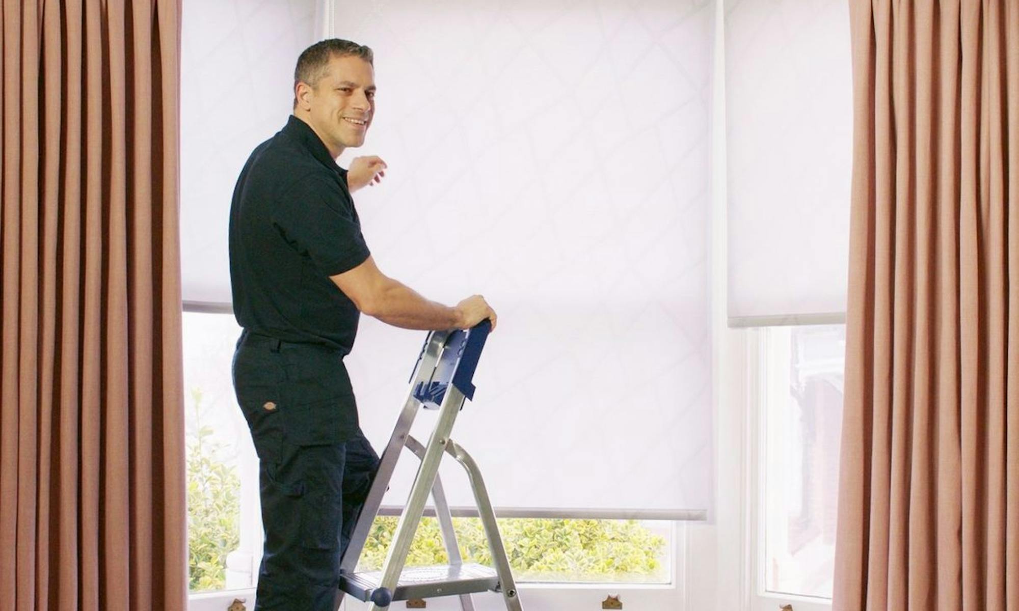Hillarys advisor wearing branded uniform, fitting white roller blinds on a window, assisting customers in their home.
