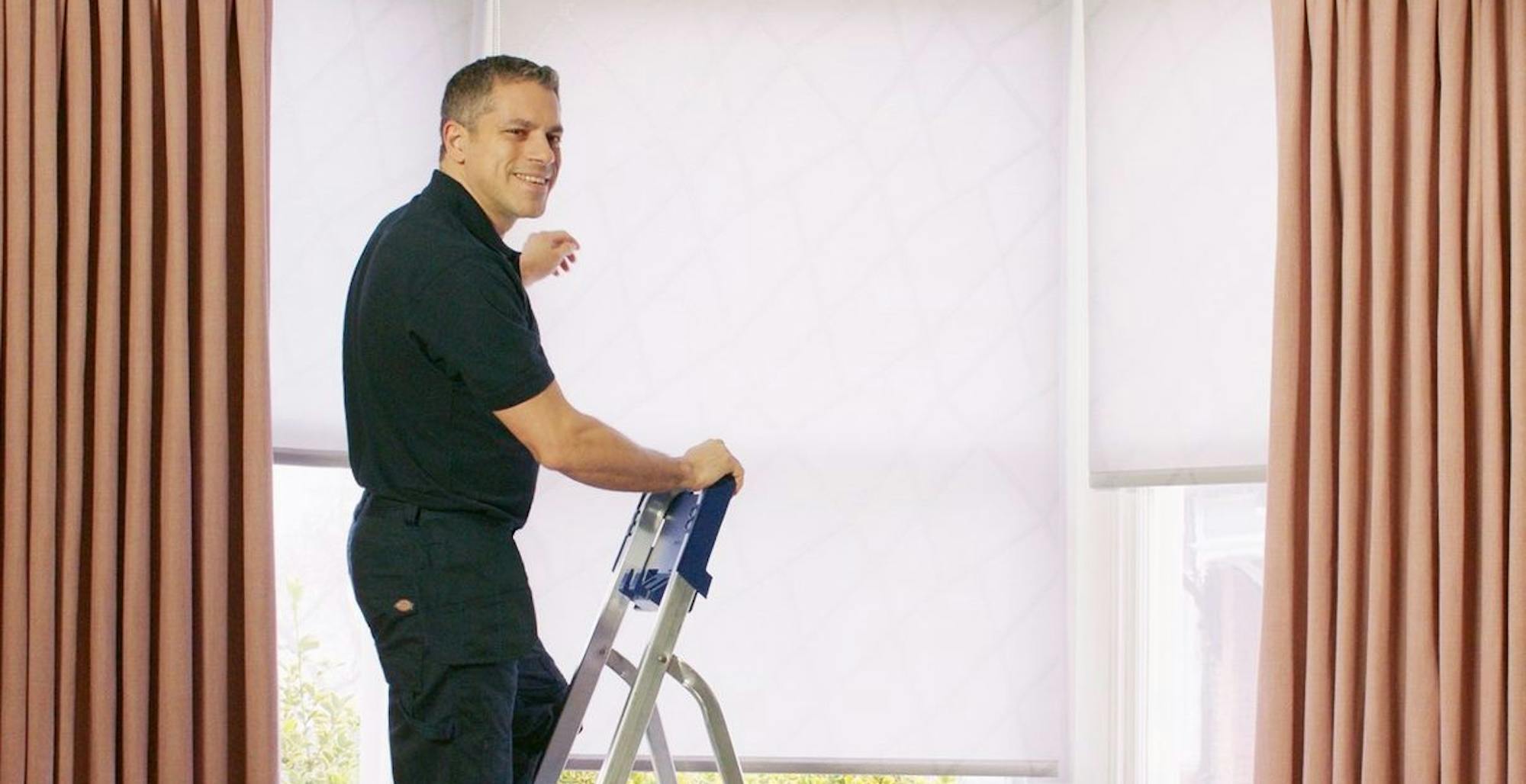 Hillarys advisor wearing branded uniform, fitting white roller blinds on a window, assisting customers in their home.