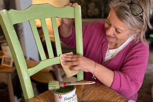 Woman smiling while sanding a green wooden chair, with a paintbrush and can of Frenchic paint on the table, preparing for a furniture makeover.