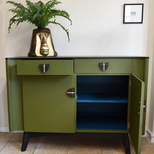 Olive green sideboard with black top and legs, featuring unique gold and black handles, open shelving, and a gold face planter with a fern on top.