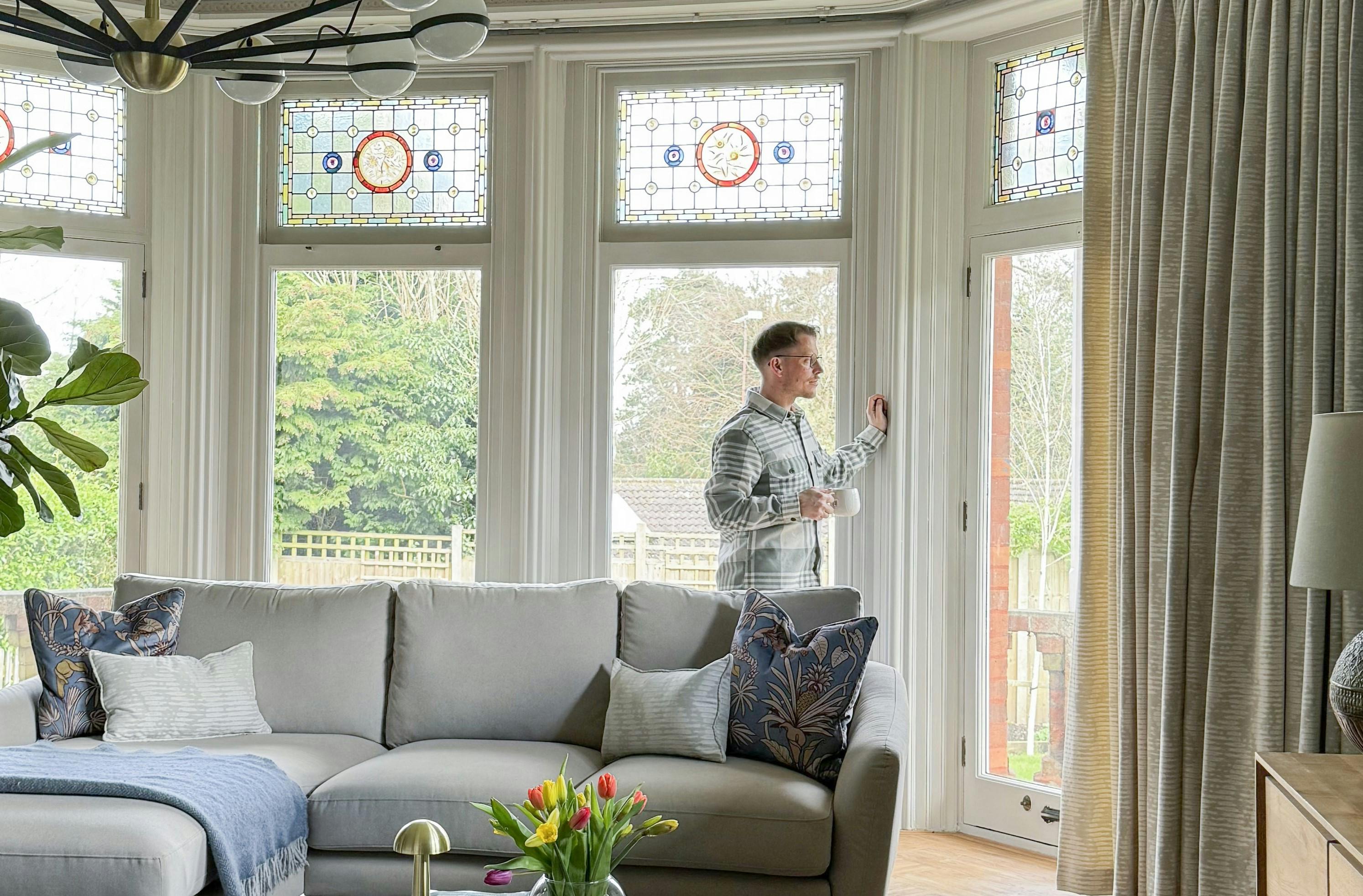 Man standing by tall windows with stained glass panels, holding a mug in a bright living room featuring a grey sofa, flowers, and cozy decor.