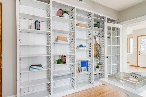 White built-in shelves with books, plants, and decor. Glass door on the right, light wood floor, and a tray on a table in the foreground.