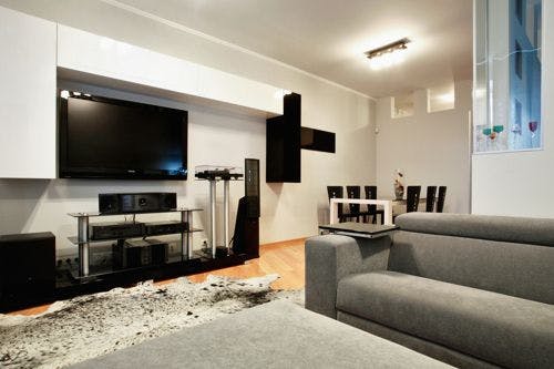 Modern living room with a large TV, gray sofa, black and white decor, and a dining area in the background with a table and chairs.