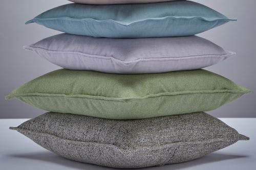 A stack of five pillows in various colors: blue, light purple, green, and two shades of gray, arranged on a white surface.