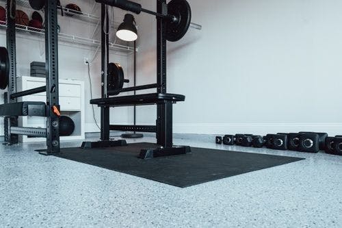 Home gym with a weight bench, barbell rack, and dumbbells on a speckled floor. A shelf with equipment and a lamp are in the background.