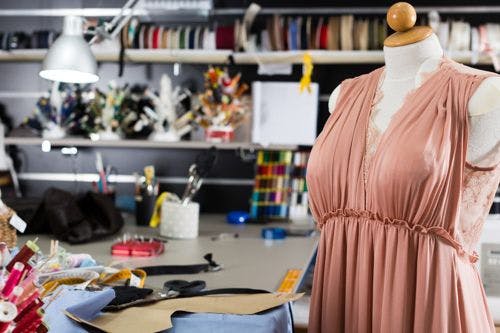 Sewing studio with a dress on a mannequin, shelves of fabric and tools, and a workspace filled with sewing materials and a lamp.
