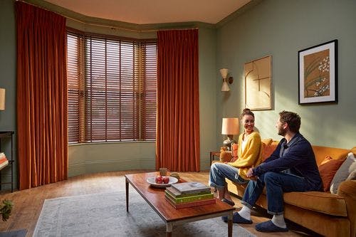 A cozy living room with green walls, orange curtains, a couple sitting on a sofa, and a wooden coffee table with books and decor.