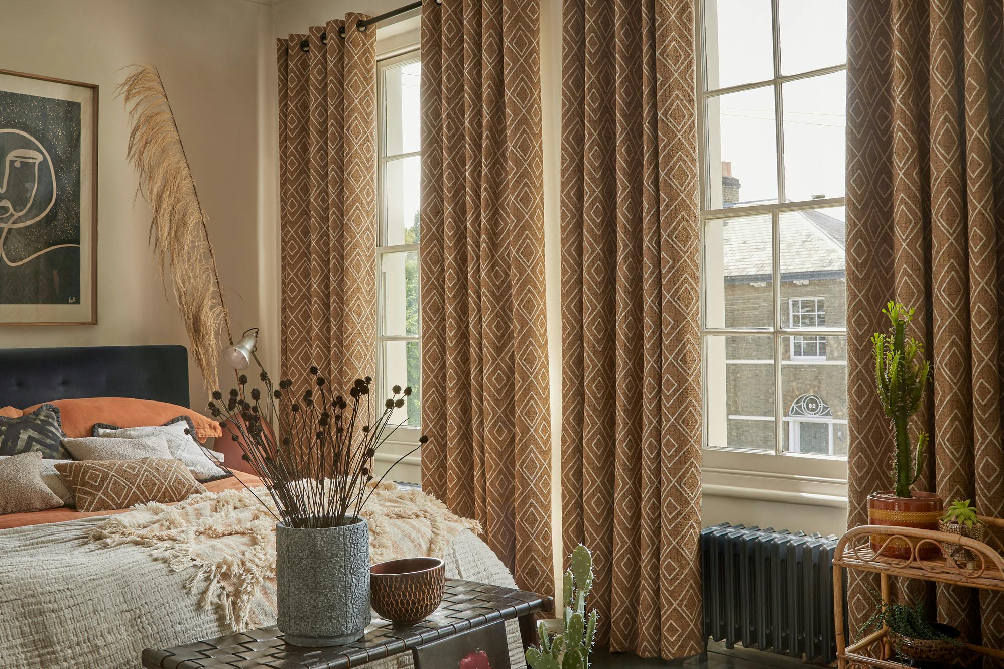Taupe bedroom with two adjacent tall windows dressed with mid cinnamon curtains with a white diamond pattern, open, bed with turmeric cover and textured throw at foor in pale beige, cushions on bed in creams, charcoal and one matching curtains, pampas by bed in base, wood and weave bench at bed end with grey vase of dried dark flowers, cactus on books on floor. 