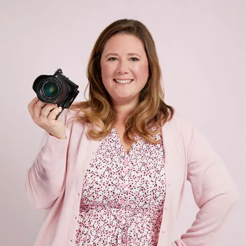 Smiling woman in a pink dress and cardigan holding a camera, standing against a light pink background, ready for a photography session.
