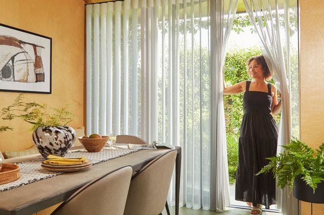 A woman entering dining room through a patio door that has sheer white diffused blinds on them.