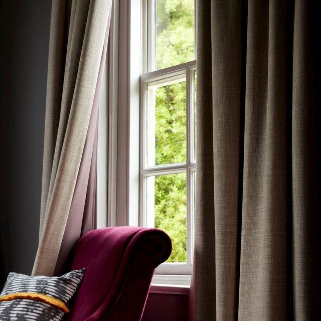 Close up of a window with light brown curtains on it and a red armchair with a cushion on top of it next to the window