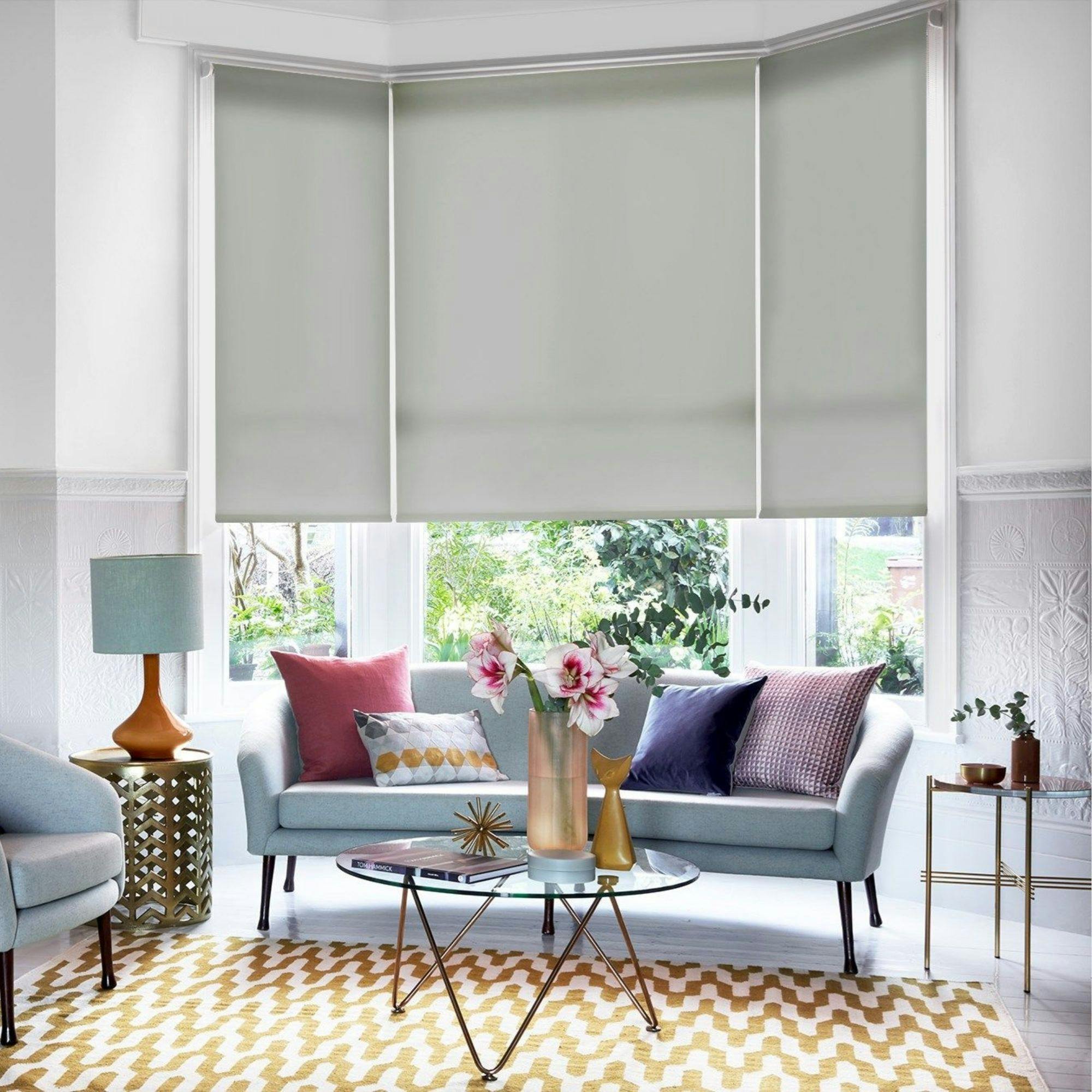 Soft‑grey blackout roller blinds fitted to a bay window in a colourful living room with pastel sofa and mustard rug.