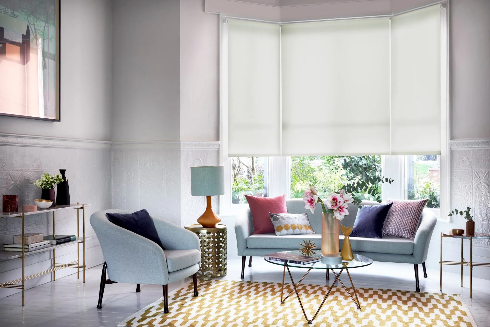 Three white roller blinds fitted to a bay window in a pastel living room with pale-blue sofa, gold-white geometric rug and glass coffee table.