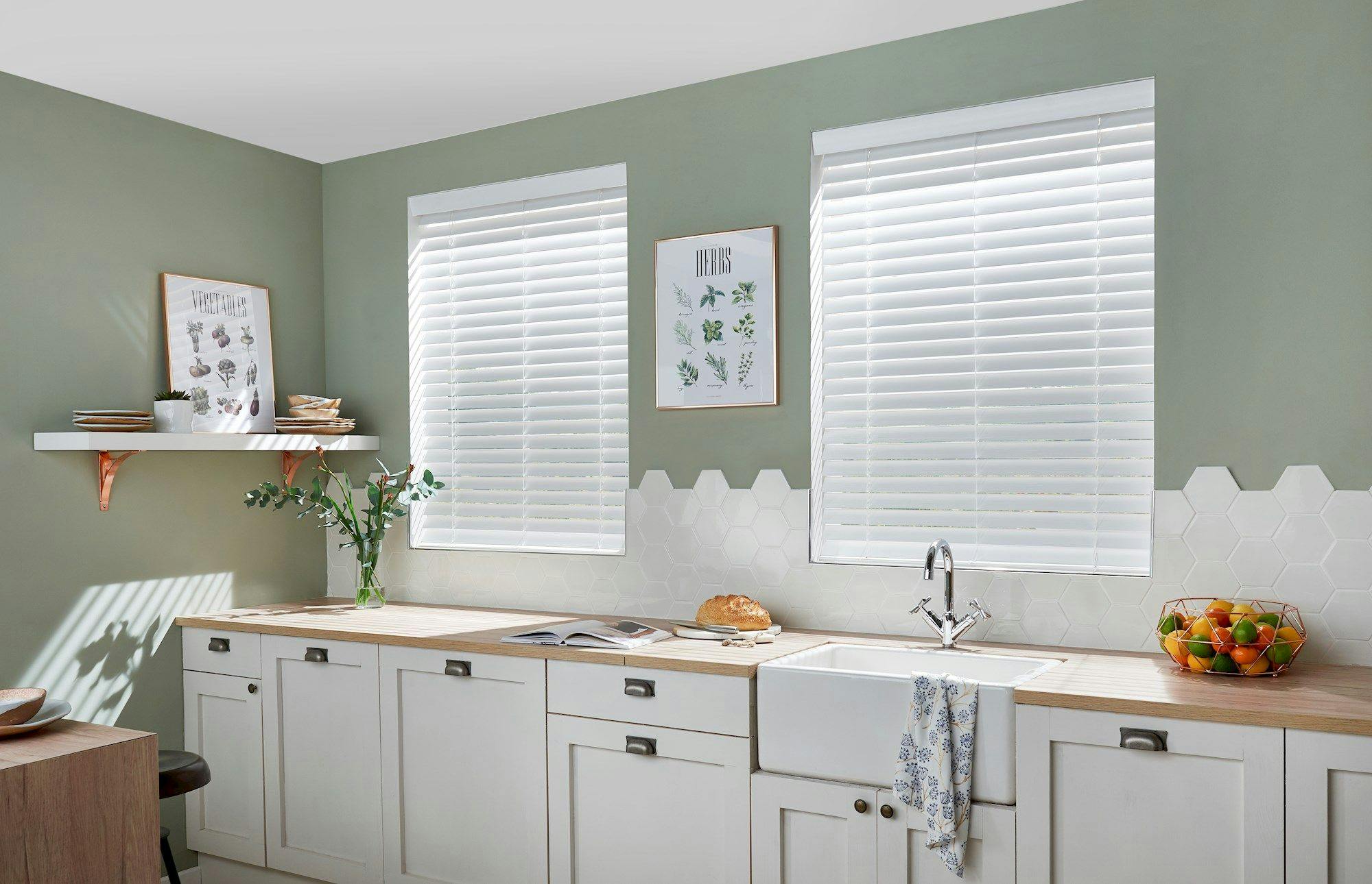 Pair of white faux‑wood venetian blinds on kitchen windows above a farmhouse sink and pale green walls.