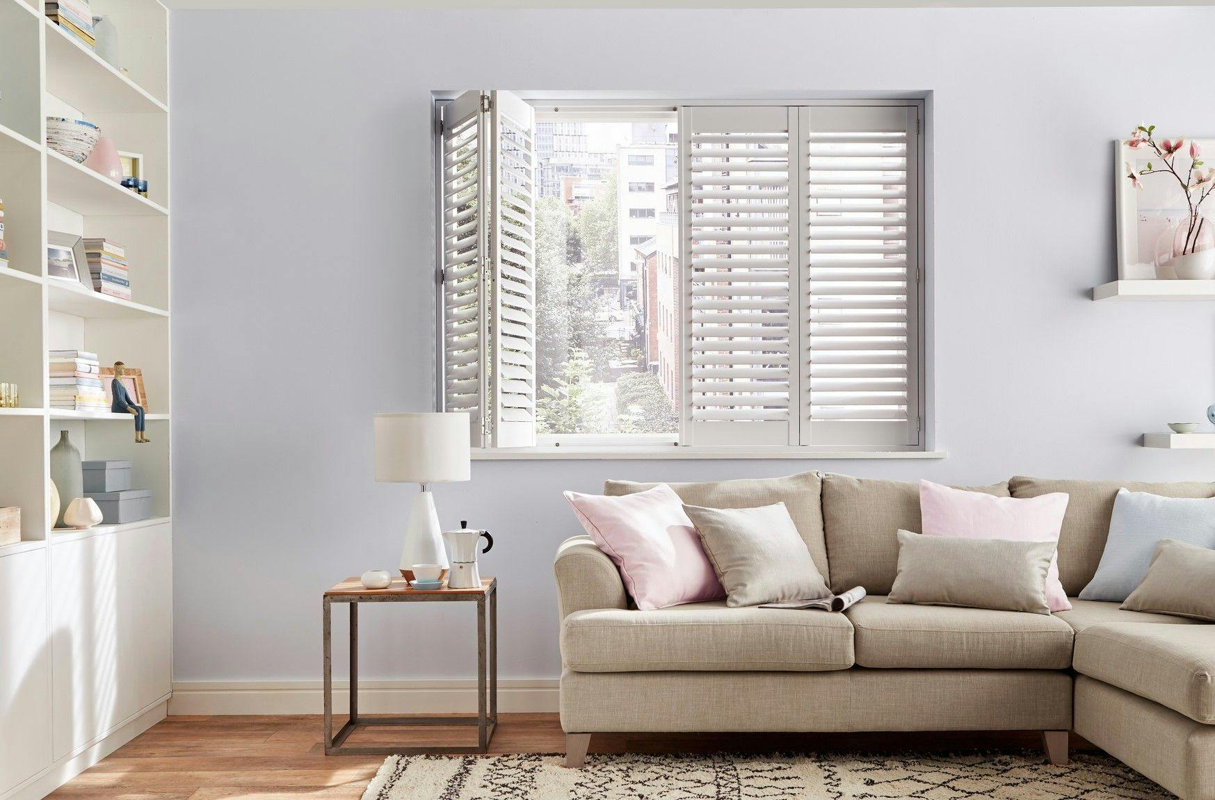 Soft-grey plantation shutters on a living-room window; one panel is open, revealing a city-street view behind a beige corner sofa with pastel cushions.