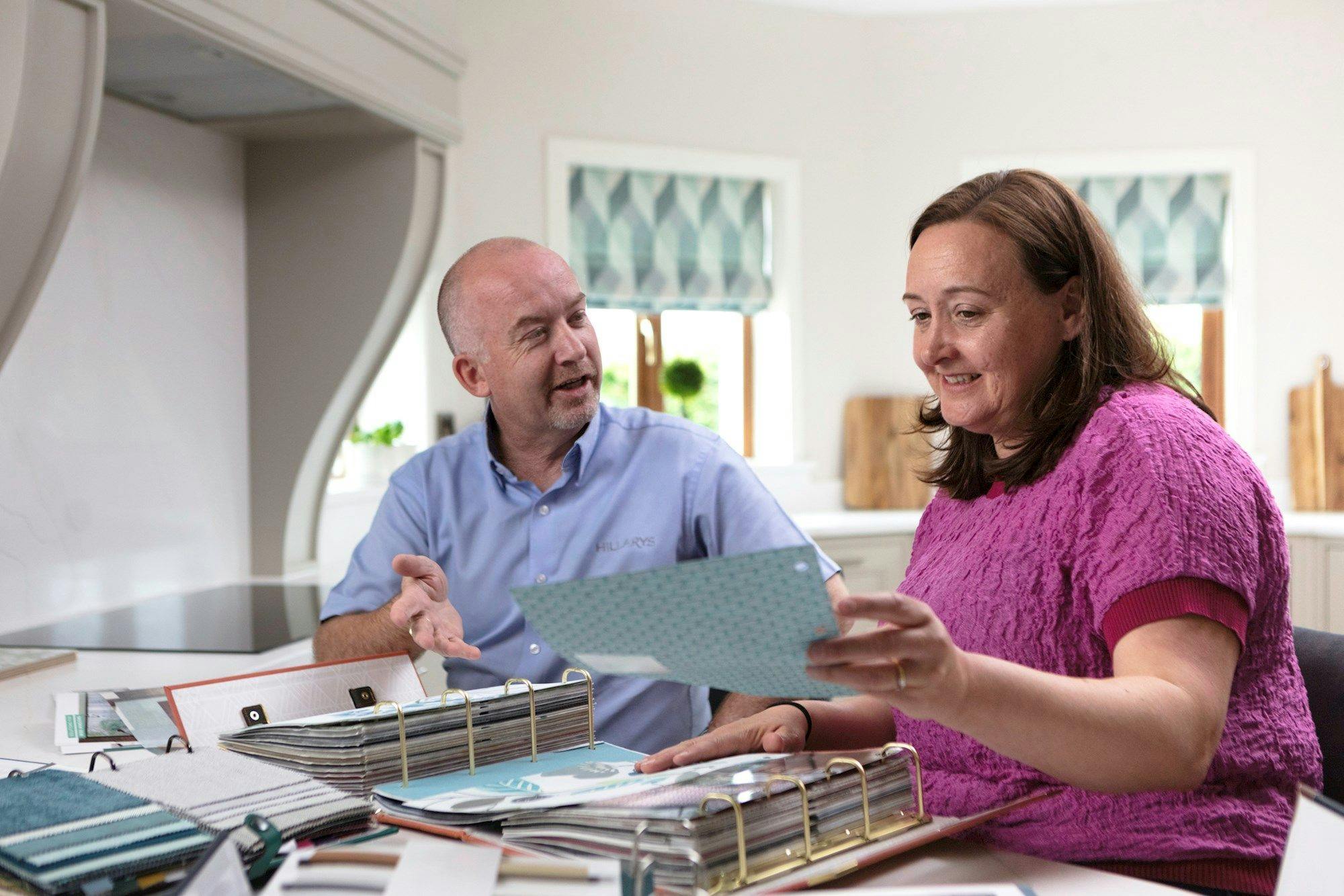 Advisor and female customer compare a teal fabric swatch from a binder at a kitchen table; matching patterned Roman blinds hang on the windows behind them.