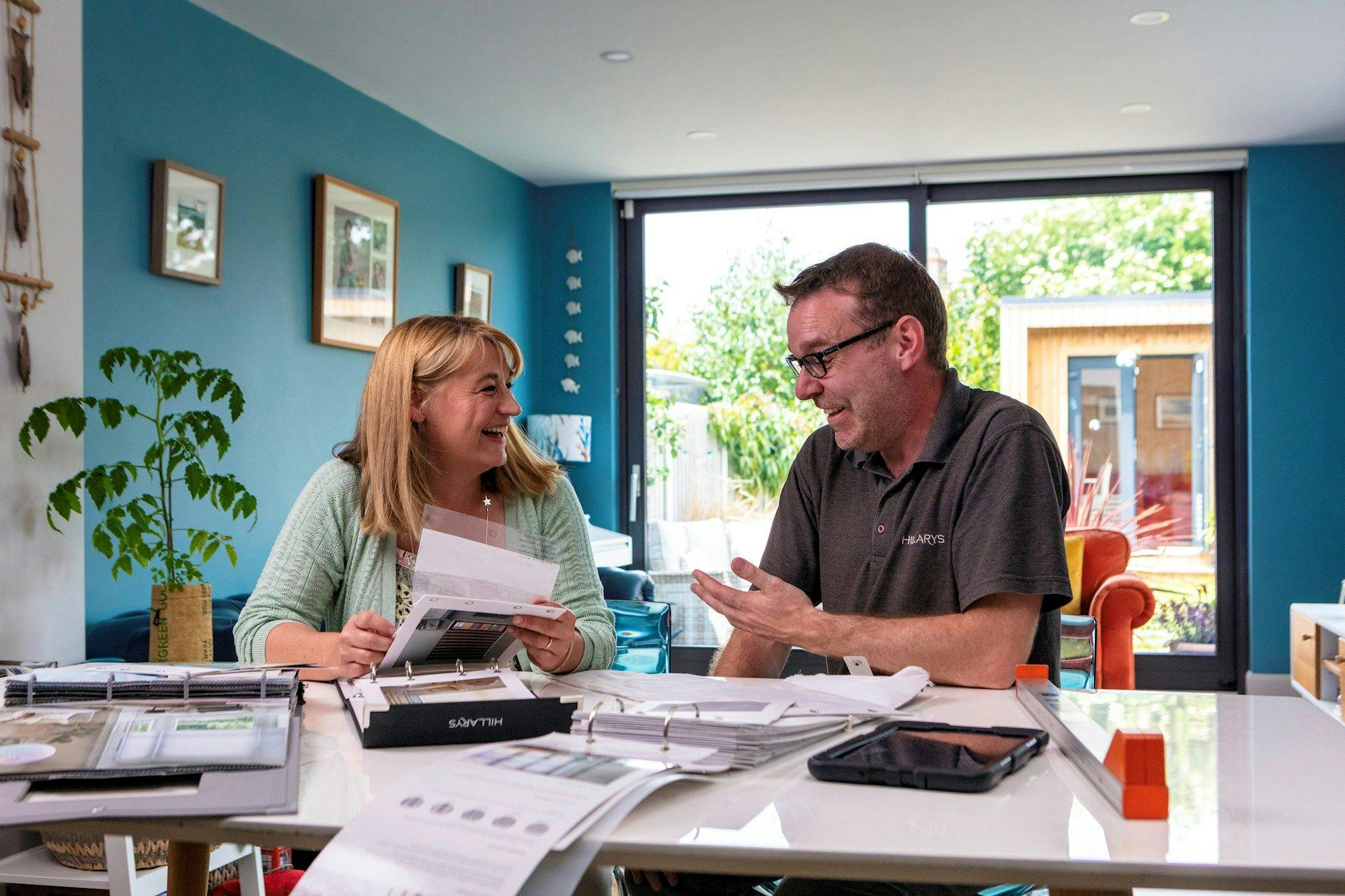 Customer and advisor laugh together during a blinds-and-curtains consultation, with multiple swatch books open on the dining-room table.