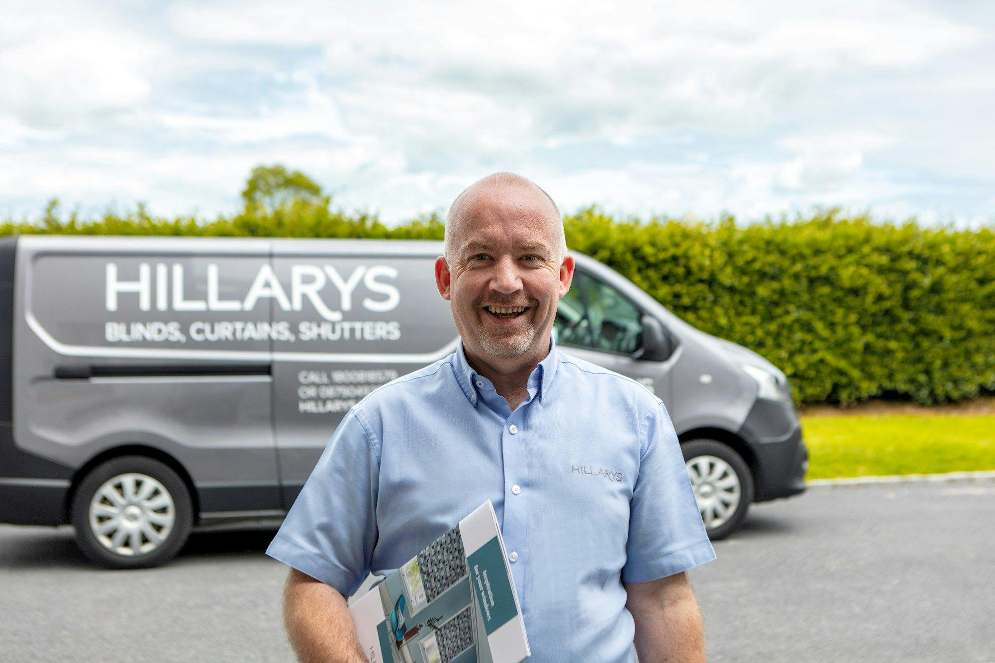 Smiling Hillarys advisor holding fabric sample folders, with a dark-grey branded van blurred in the background.