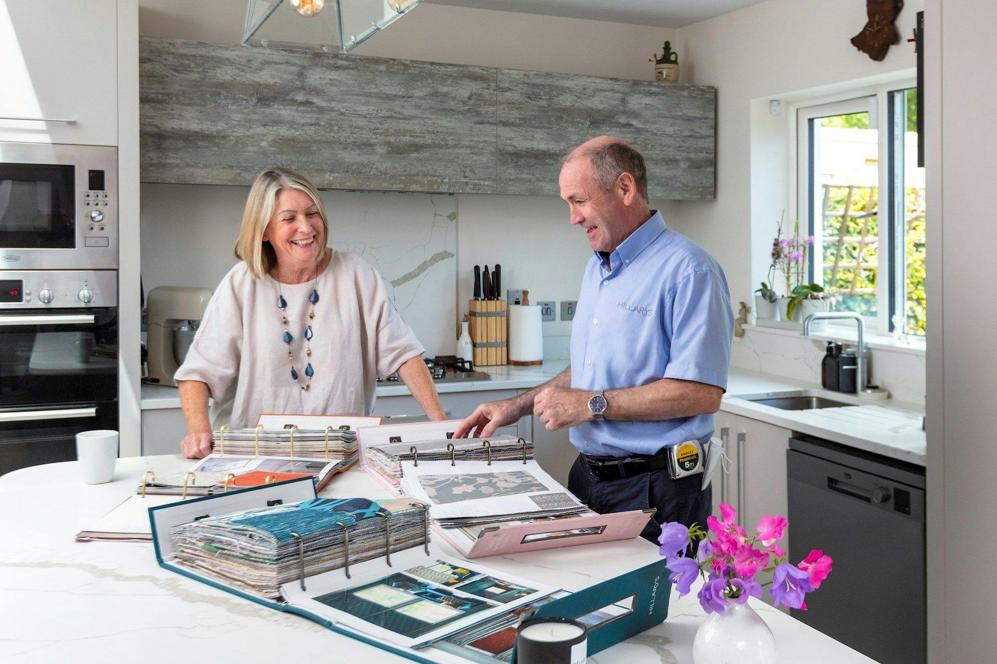 Advisor points to a page in a fabric-swatch binder on a white kitchen island while the homeowner smiles; several open sample books lie around them.