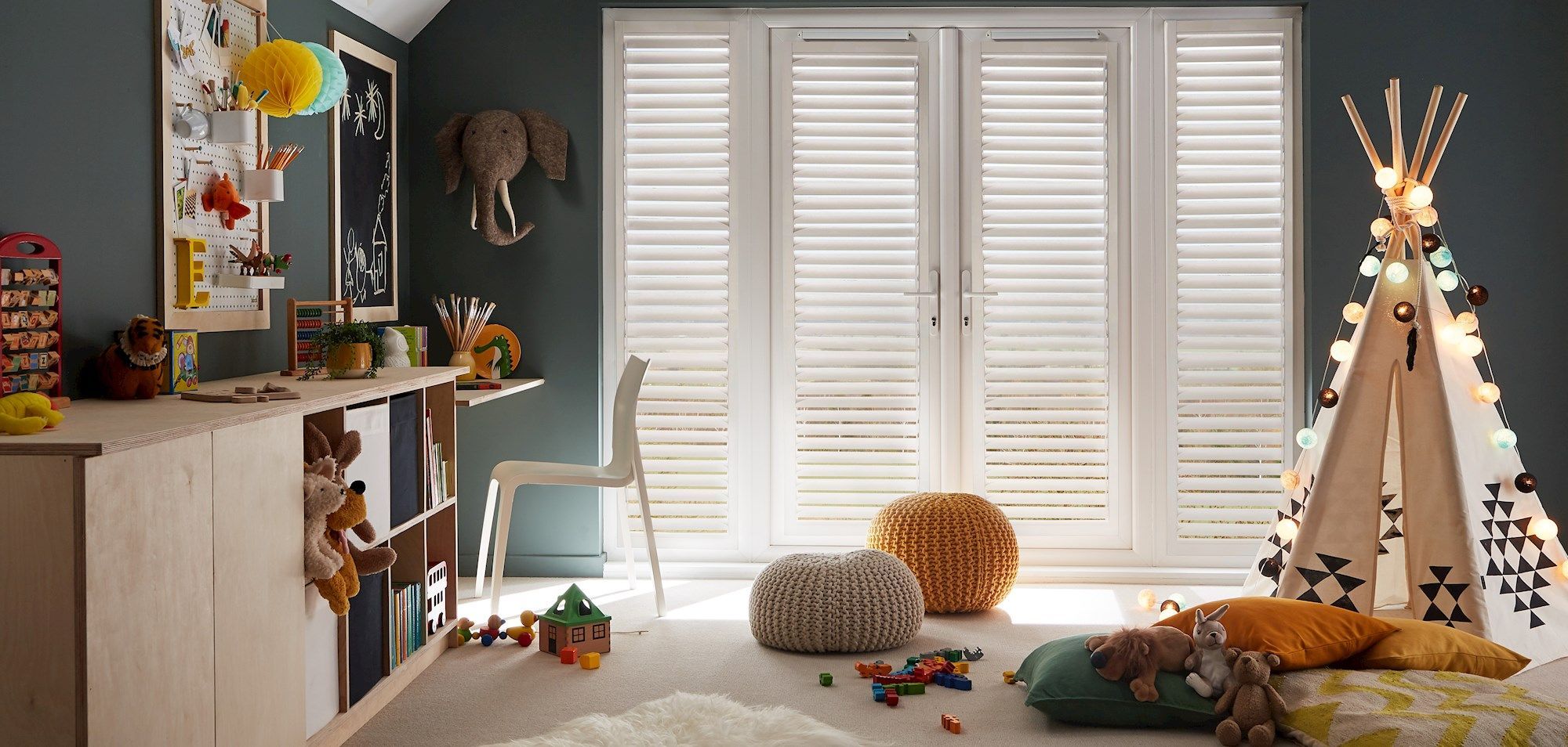 Children’s playroom featuring white shutters on French doors, a teepee draped with string lights, wooden storage, pouffes and scattered toys.