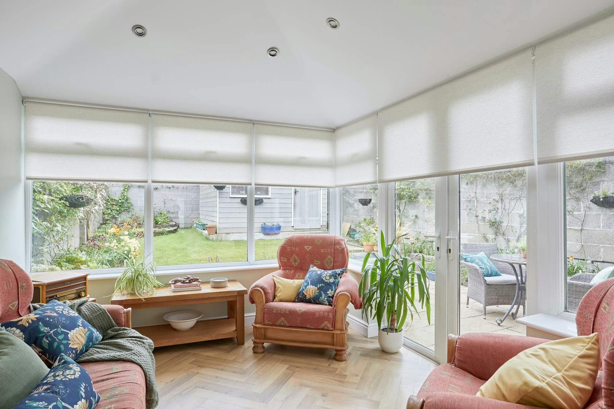 A conservatory with parquet floor, wooden-framed upholstered terracotta chairs, with sheer off-white Roller blinds partially lowered.