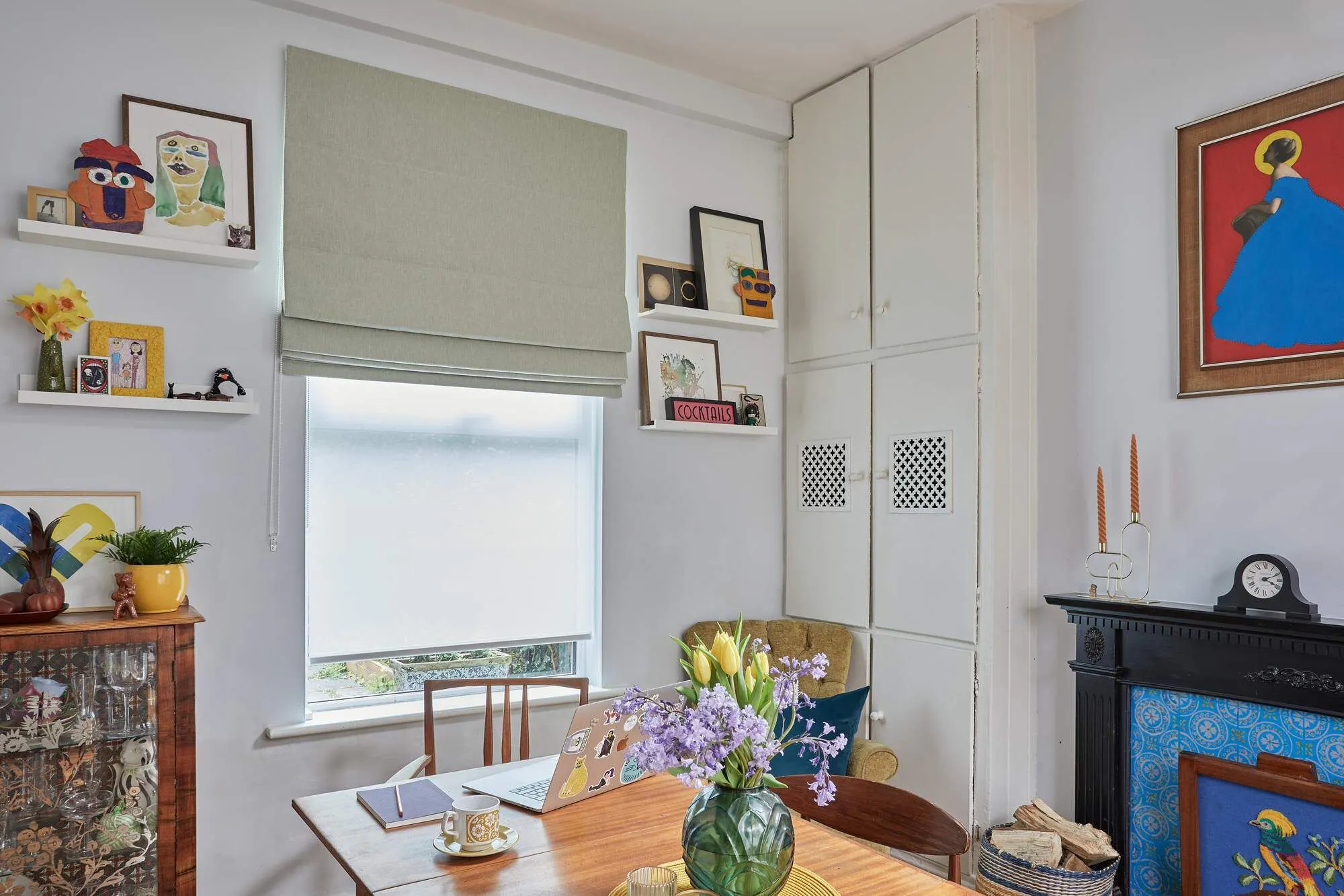 A dining room with wooden floor, fireplace with decorative screen of a bird, white walls, and a window dressed in a pale green Roman blind with a sheer white roller blind underneath.